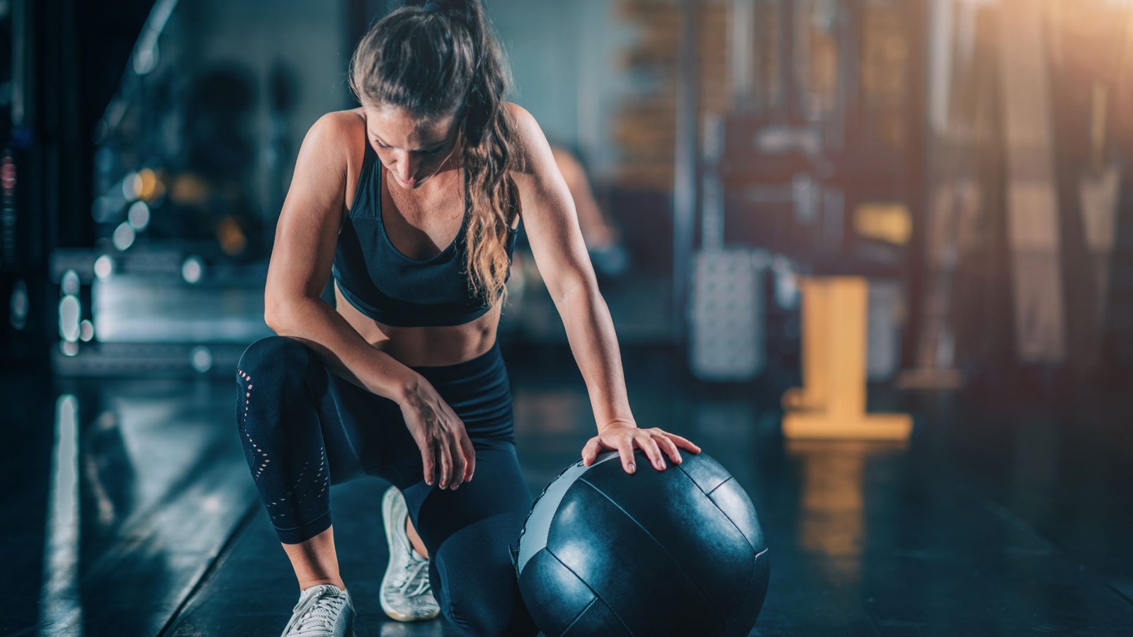 Woman in athletic wear kneels beside a medicine ball in a gym.