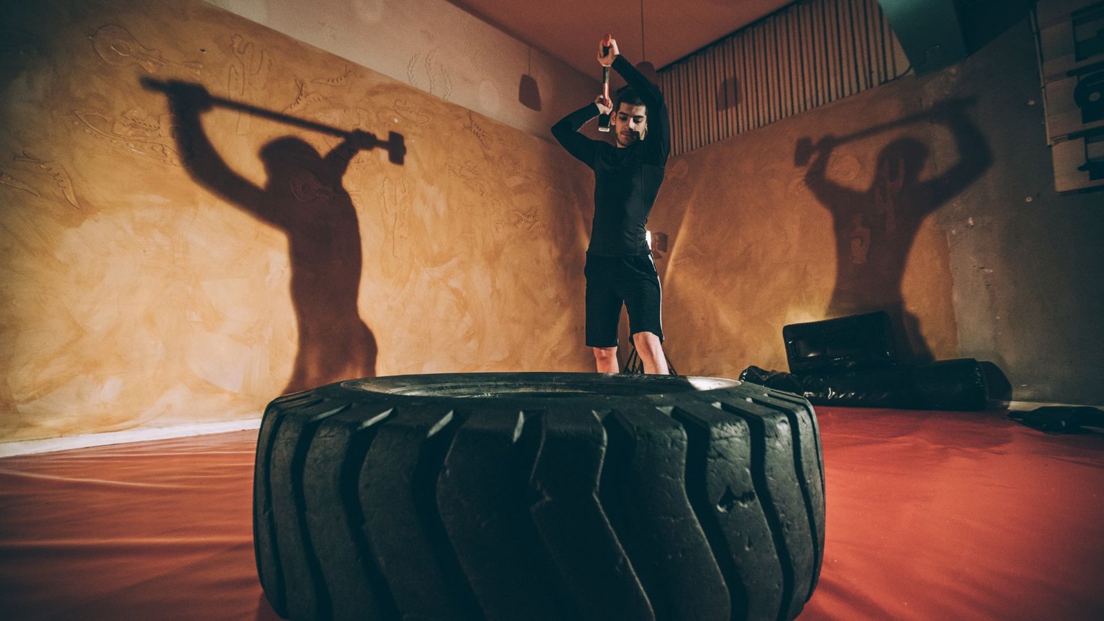 Person in black athletic wear swings a hammer over a giant tire in a dim gym, with large wall shadows.