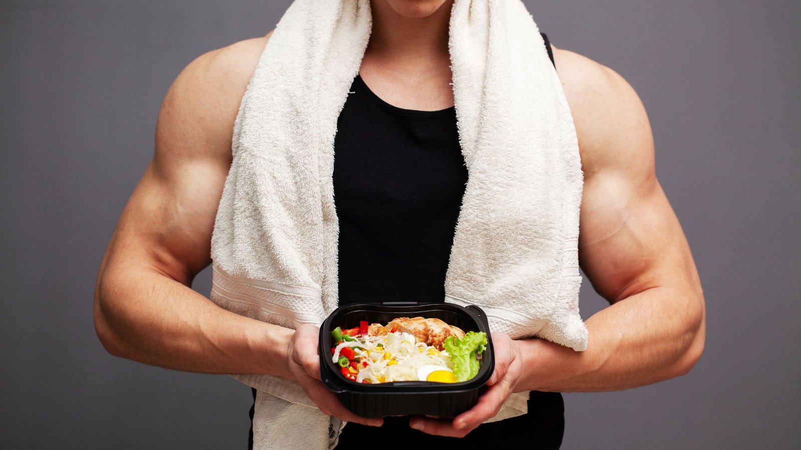 Person in a black tank top with a towel draped over shoulders holding a black takeout container with colorful salad and chicken.